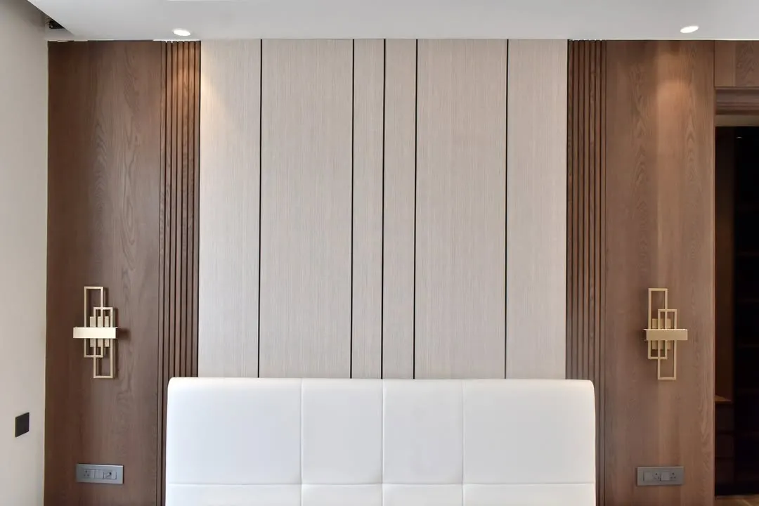 This integrated bedroom wall design combines function and elegance. A plush white headboard is set against a backdrop of light and dark wood panels, featuring modern fluted details and stylish gold sconces for ambient lighting.