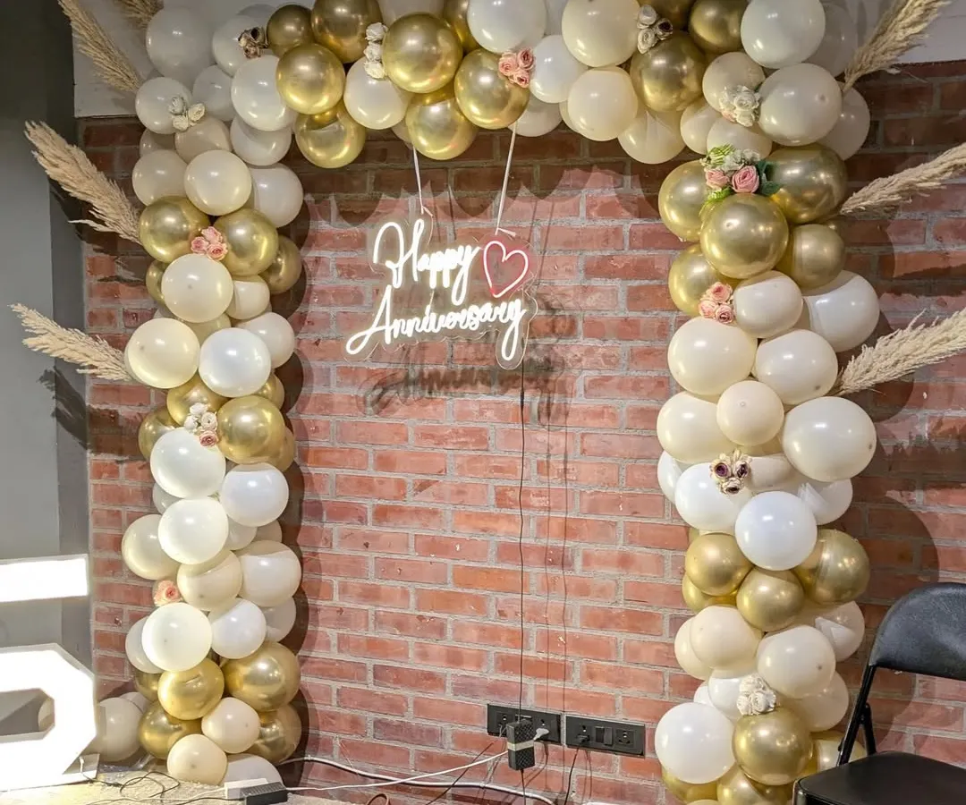 I design elegant backdrops for milestone celebrations like this 25th anniversary. A custom balloon arch in a sophisticated gold and white palette, paired with a 'Happy Anniversary' neon sign, creates a beautiful photo corner.