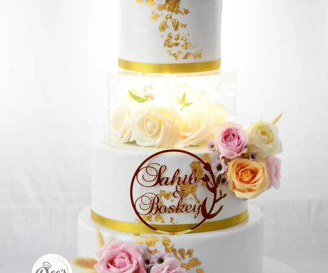 A wedding cake should be as timeless as your love story. This four-tier beauty features an illuminated acrylic section filled with roses, delicate sugar flowers, and touches of edible gold leaf for a truly showstopping centerpiece.