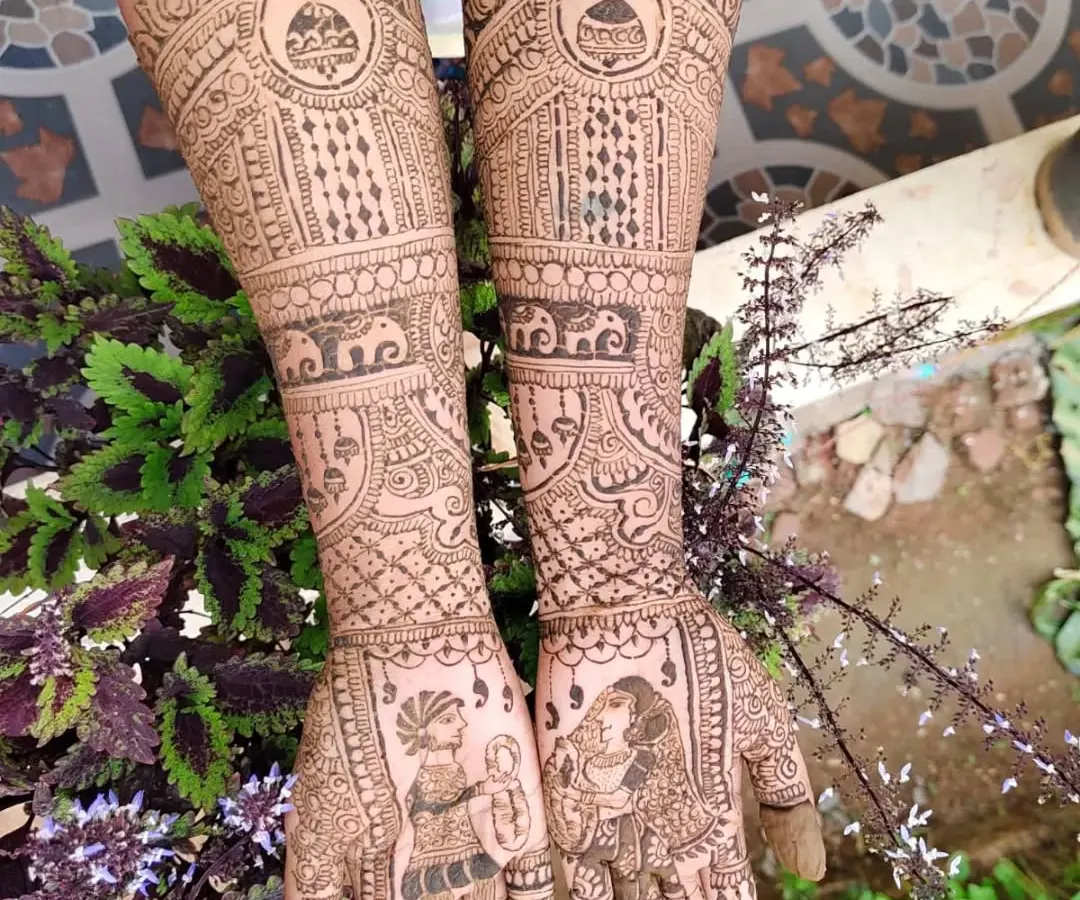 A full bridal mehendi design that goes up to the elbows, showcasing traditional Indian artistry. The palms feature classic dulha-dulhan (bride and groom) figures, while the arms are filled with intricate patterns, including elephants, kalash motifs, and delicate jaal work, all done with natural henna.