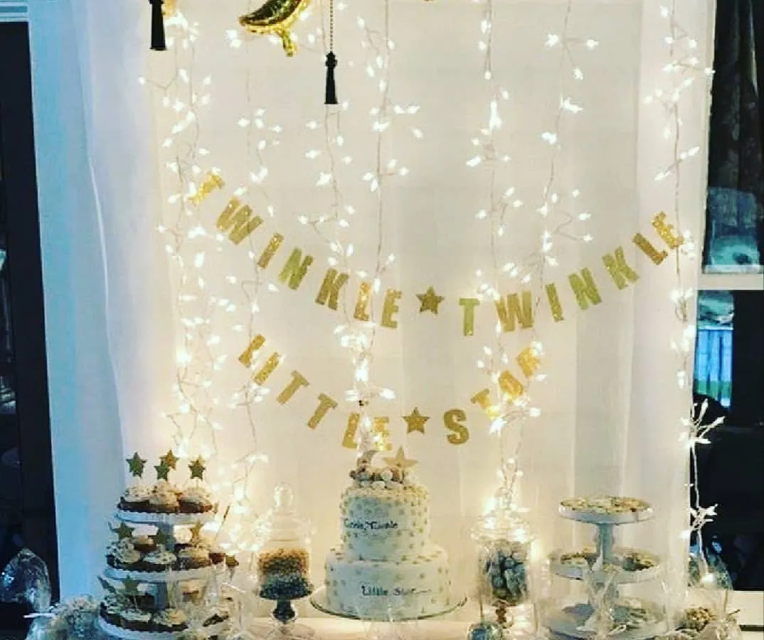 The 'Twinkle Twinkle Little Star' theme set up for a baby shower dessert table. This decoration features a curtain of warm fairy lights, a golden banner, and foil moon and star balloons, making the cake and treats stand out.