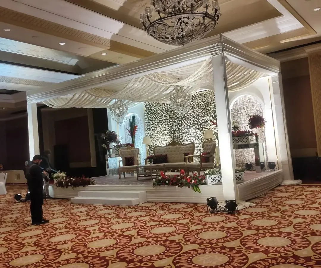 An elegant and grand indoor wedding stage setup within a banquet hall. The design features a pristine white mandap structure, a dense floral backdrop of white flowers, and luxurious seating, all under a classic crystal chandelier.