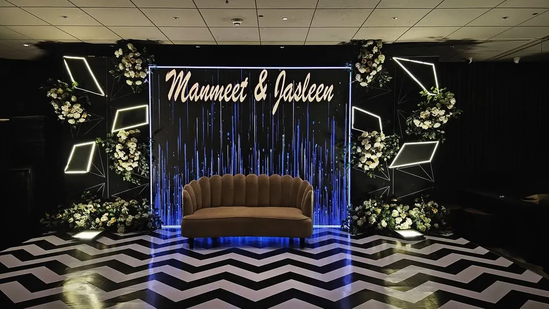 A chic and modern reception stage for Manmeet and Jasleen. The design features a dramatic black and white chevron floor, custom neon-style lighting, and elegant white floral arrangements for a high-impact, contemporary look.