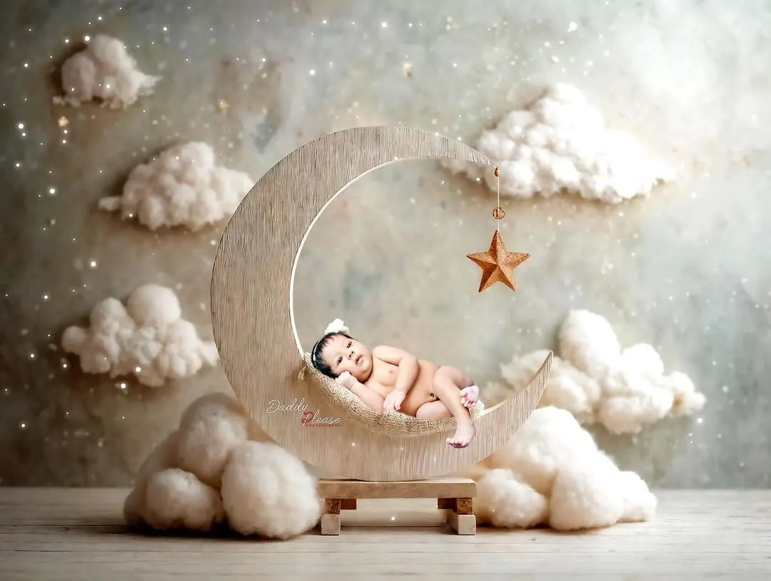 A peaceful newborn portrait featuring a baby sleeping on a wooden crescent moon prop, surrounded by soft, dreamy clouds. These sessions are designed to be calm and safe for your little one.
