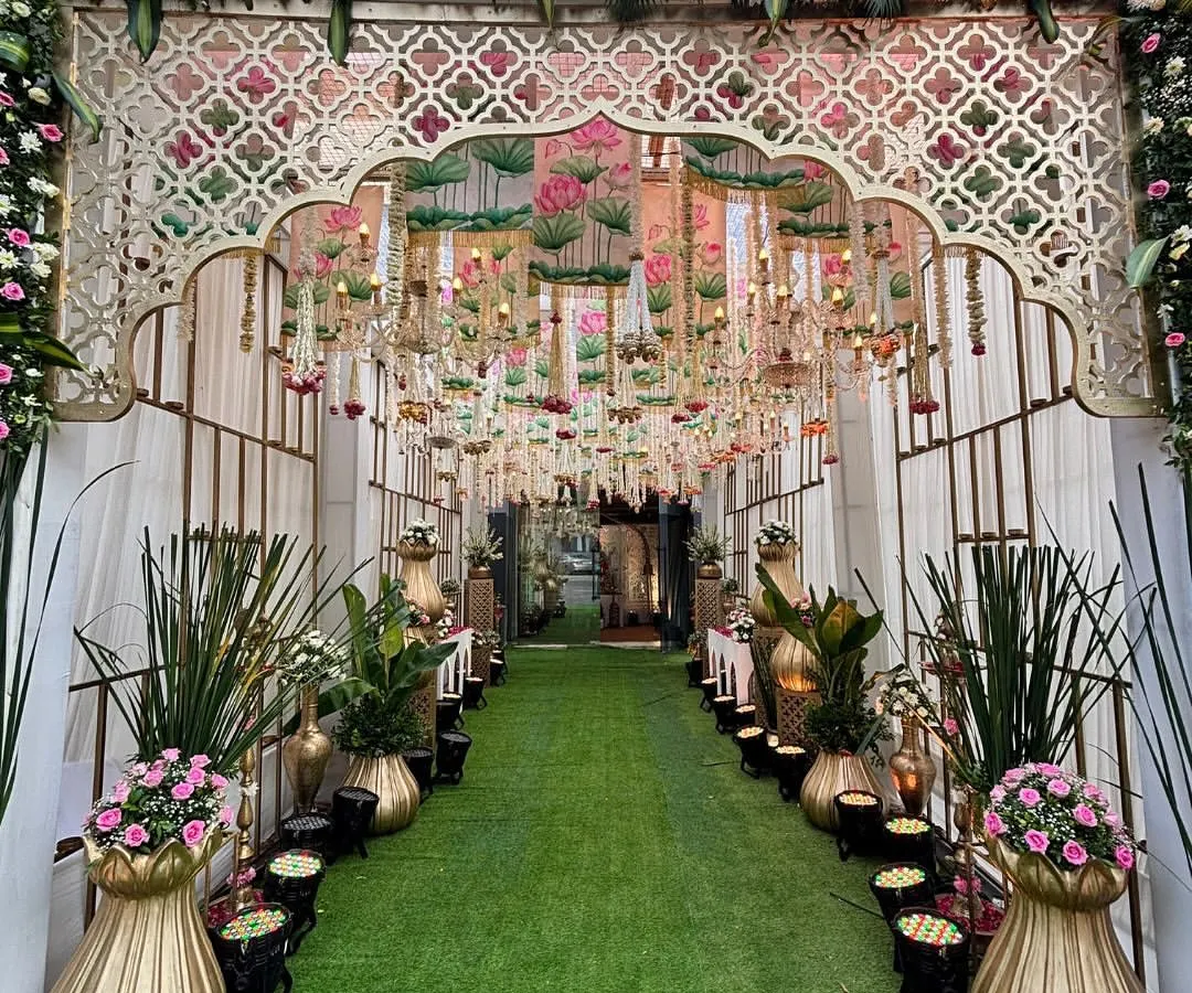 Your guests' first "wow" moment starts here. I love creating immersive entrances like this one, with a lush green carpet, grand floral arches, and a ceiling of hanging chandeliers and lotus-themed drapes to build anticipation for the celebration ahead.