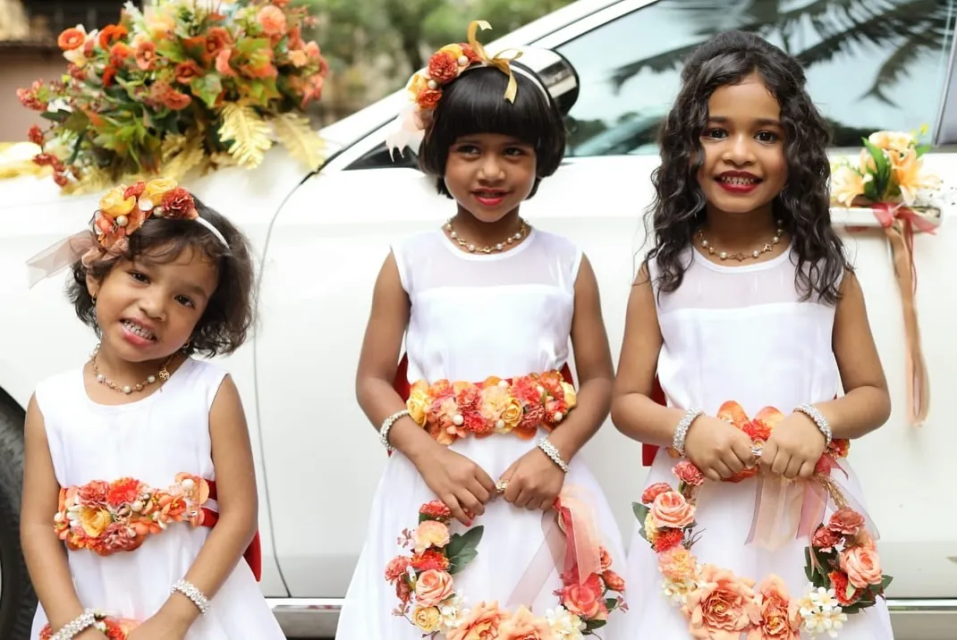 Look at these adorable flower girls. I designed their matching floral accessories, including head wreaths, waist belts, and floral hoops, using a vibrant orange and peach color palette to complement their white dresses and the decorated wedding car. These are the moments that make a wedding unforgettable.