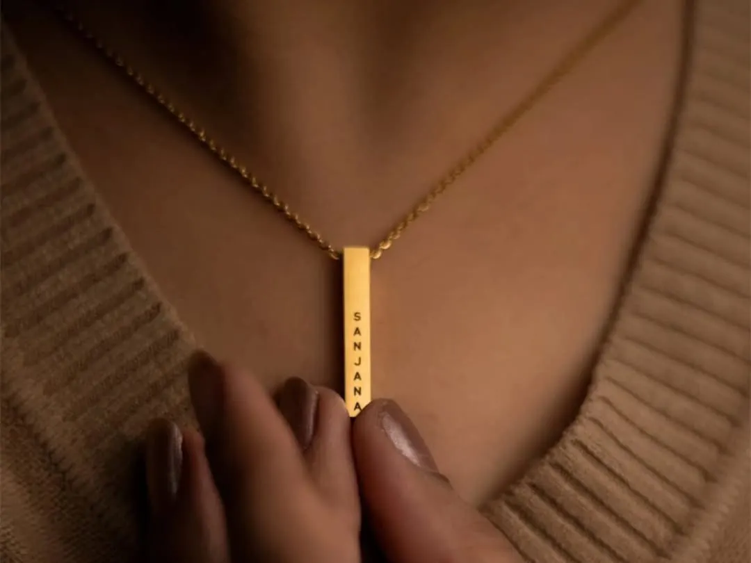 This close-up shows a gold bar pendant engraved with the name 'Sanjana'. The clean font and minimalist design make it a versatile piece, perfect for wearing alone or layering with other necklaces.