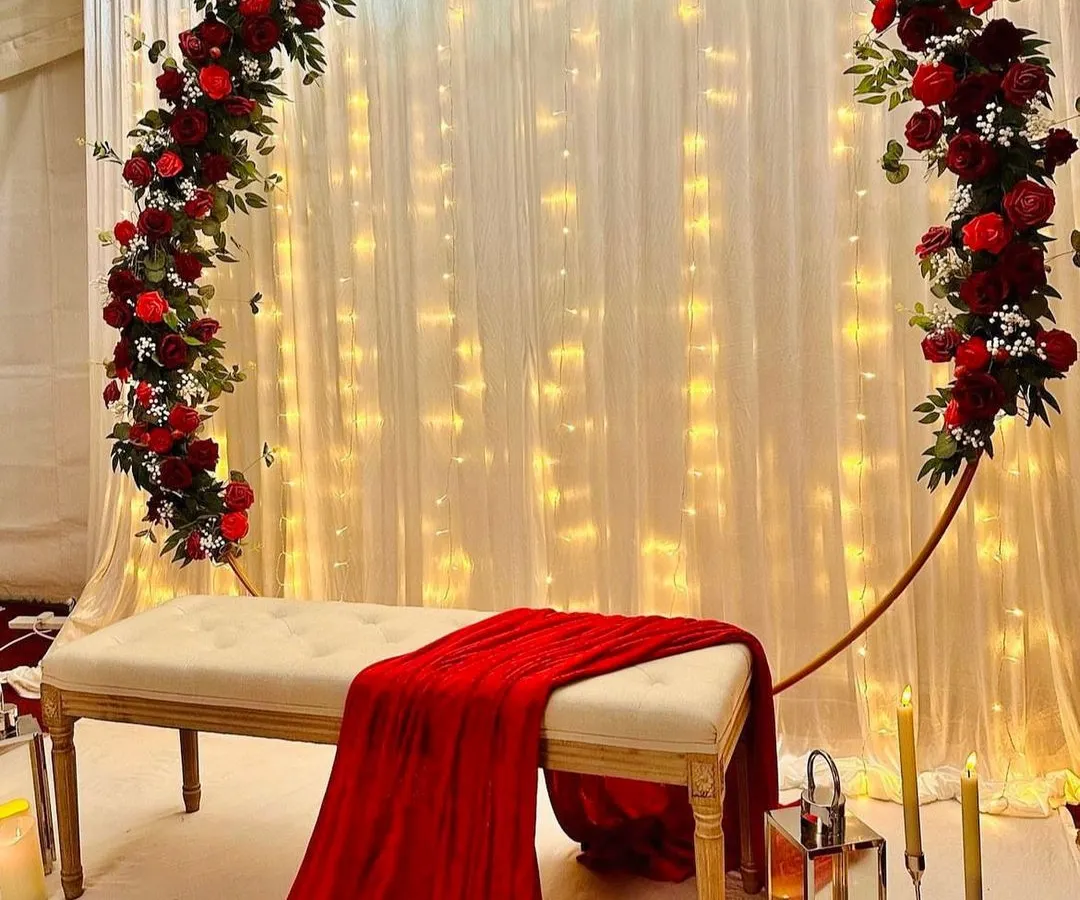 For a classic and passionate statement, you can’t go wrong with deep red roses. I designed this romantic backdrop with a full floral arch over a simple white bench, set against a curtain of warm fairy lights. This elegant stage decoration is perfect for making your engagement photos pop.