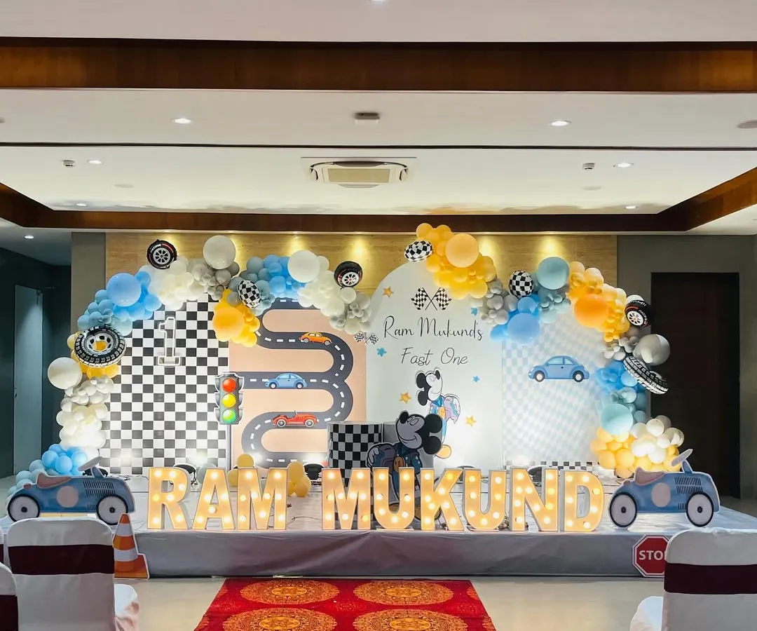 A 'Two Fast Two Curious' car-themed first birthday party. This large-scale setup features a custom printed backdrop with a racetrack, a multi-color balloon arch, car cutouts, and light-up marquee letters spelling out the birthday boy's name.
