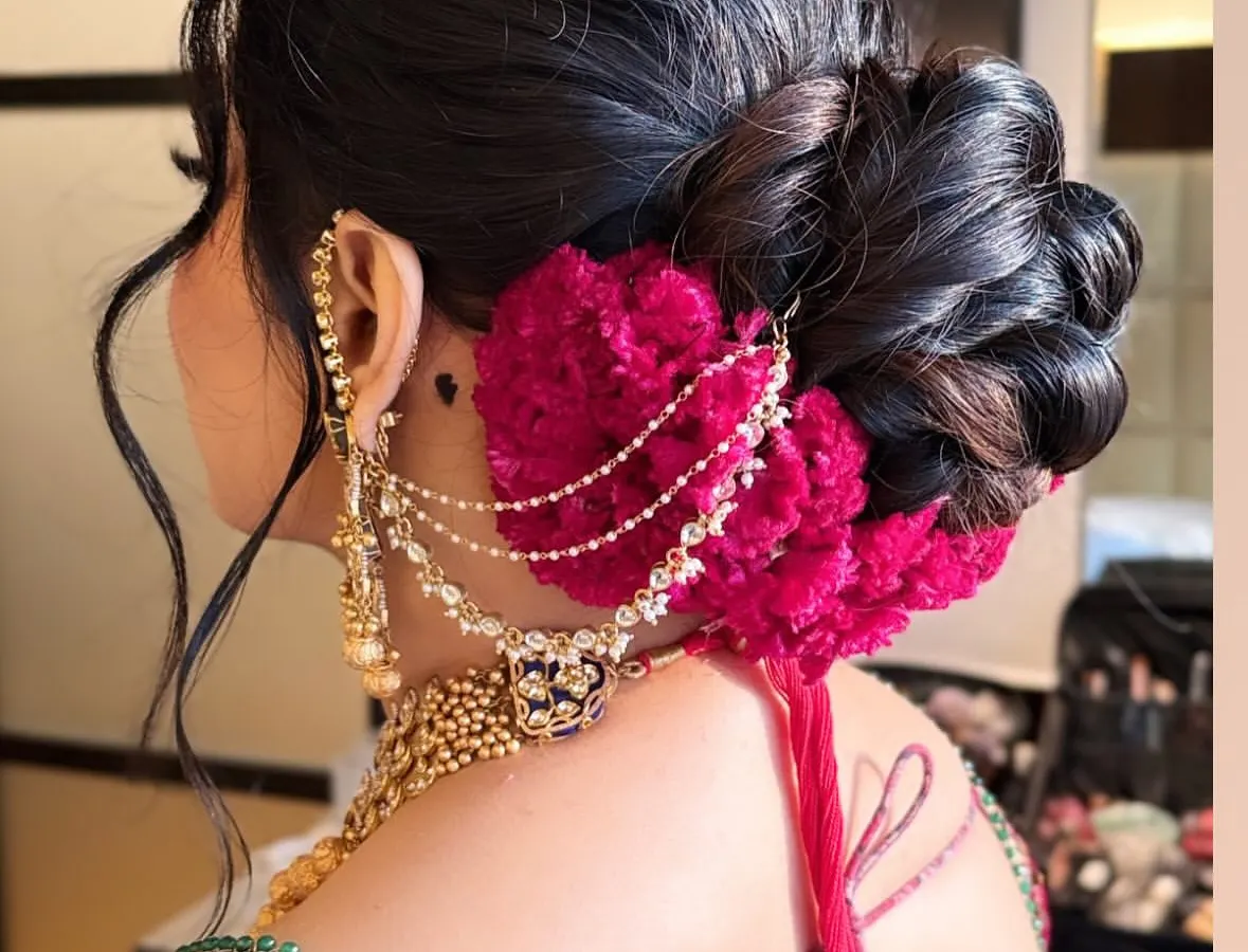 A voluminous and intricately textured bridal bun, perfect for a North Indian wedding ceremony. This style is accented with deep magenta flowers and a delicate chained accessory, creating a look that is both classic and striking from every angle.