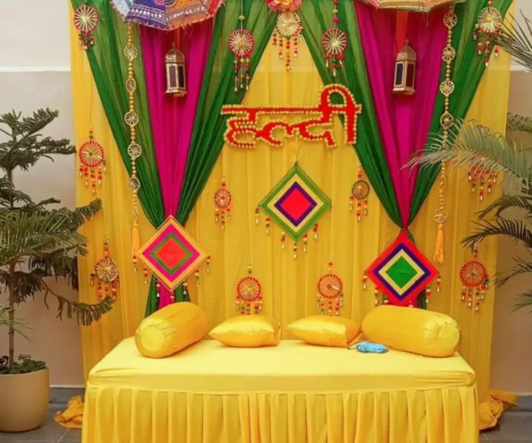 A classic Haldi ceremony backdrop designed for a home setting. This setup features a bright yellow diwan for seating, framed by vibrant yellow, pink, and green drapes, and decorated with traditional phulkari umbrellas and hanging tassels for a festive look.