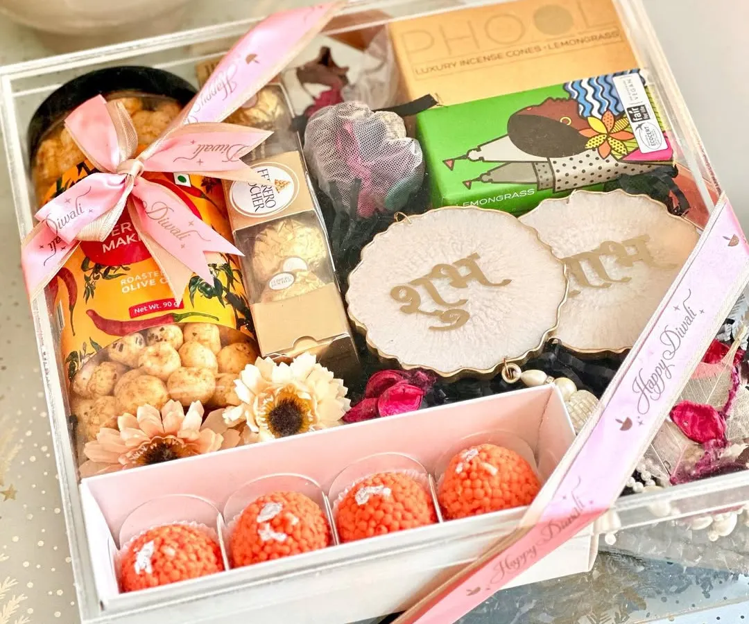 We believe a gift should be an experience. This clear acrylic box for Diwali is filled with festive joy, including traditional ladoo-shaped candles, Shubh Labh wall hangings for blessings, gourmet makhana, and rich chocolates, all tied together with a personalized ribbon.