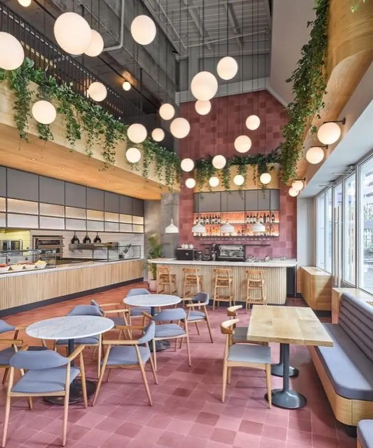 We designed this spacious, double-height cafe to feel vibrant and full of life. The design features a bold pink tiled floor, an array of spherical pendant lights for dramatic effect, and hanging greenery to soften the industrial architecture, creating an energetic and welcoming atmosphere for customers.