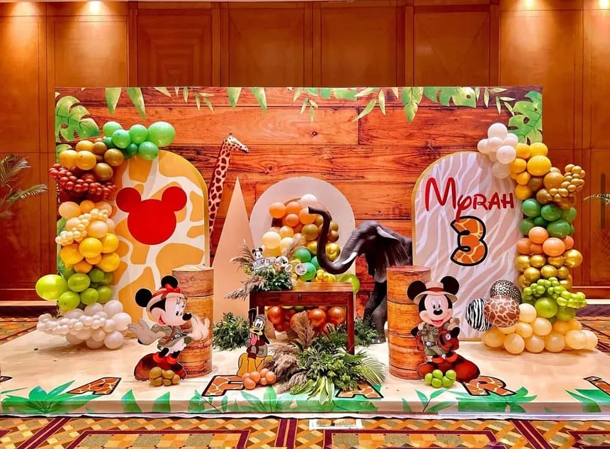 Welcome to a Mickey Mouse jungle safari adventure! This expansive backdrop brings the wild indoors with a wood-paneled wall, lush balloon garlands in earthy tones, and beloved characters like Mickey and Pluto ready for exploration. It's a perfect example of how I transform a space into a story.