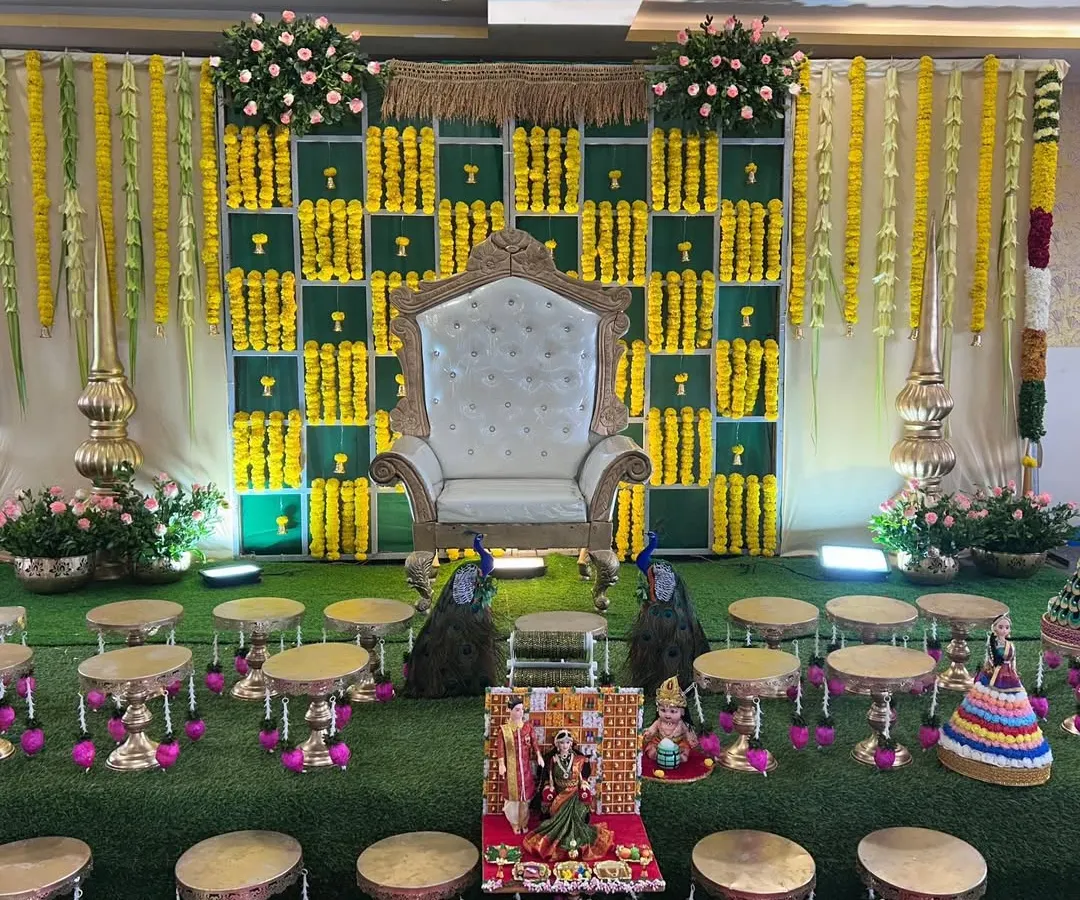 An elaborate and traditional baby shower stage, rich in cultural details. The decor includes a checkered backdrop of green panels and yellow marigolds, a regal white throne, hanging floral garlands, and brass accents, all set on a green turf for an authentic feel.