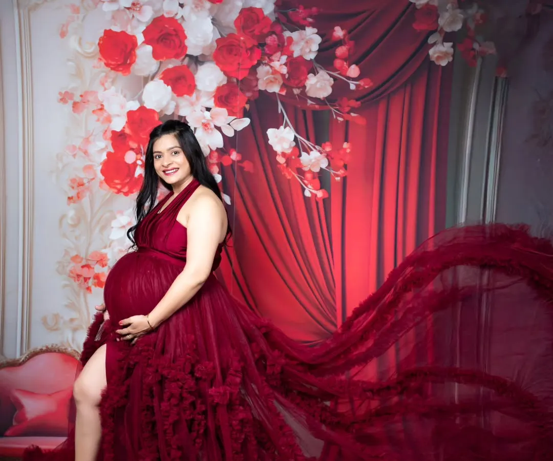 A glamorous maternity portrait of a mother-to-be in a stunning, floor-sweeping red gown against a dramatic floral backdrop.