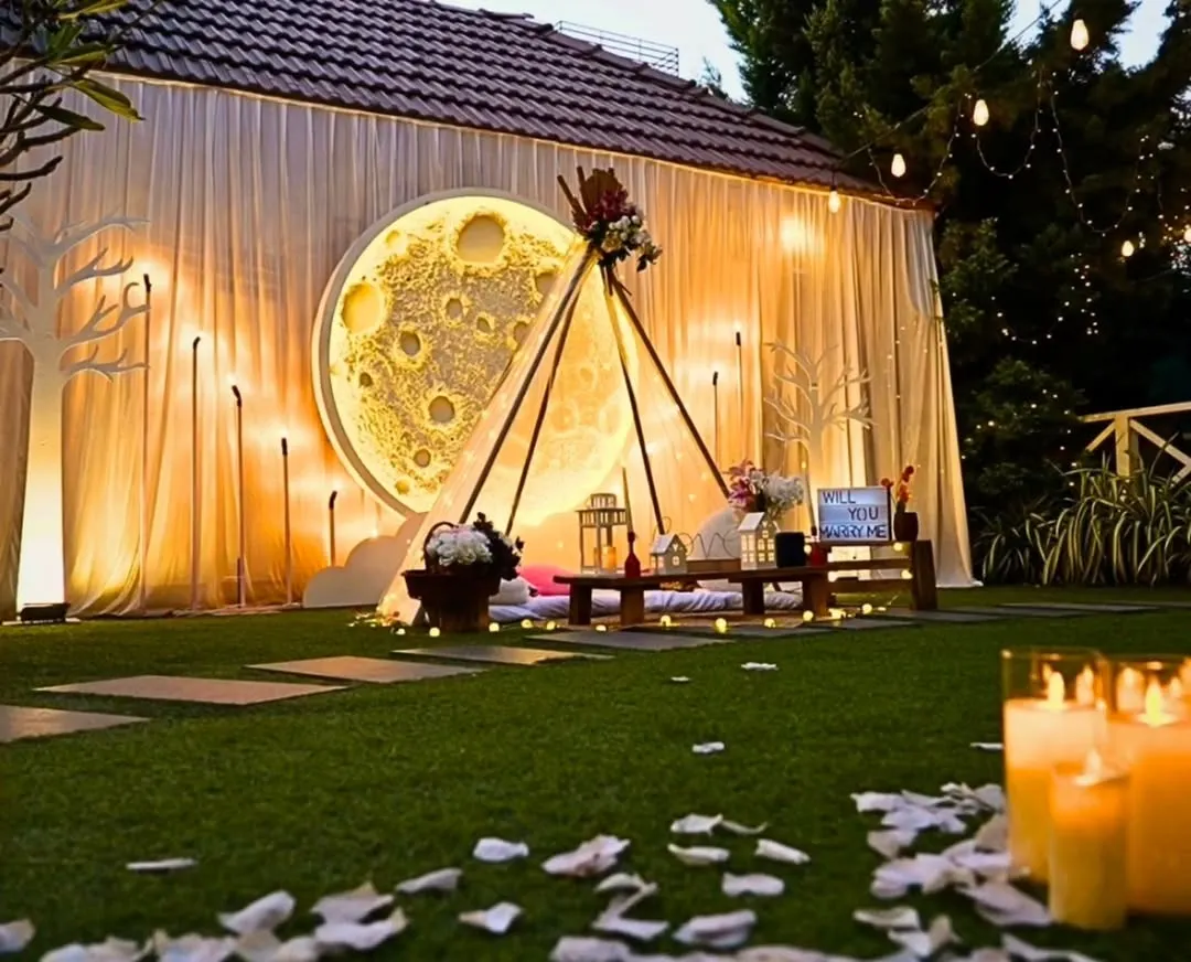 This "Love you to the moon and back" themed setup is perfect for a magical surprise. It features a large, glowing moon backdrop, a cozy teepee for two, and a pathway of scattered petals and candles, all set in a private garden for an unforgettable evening.