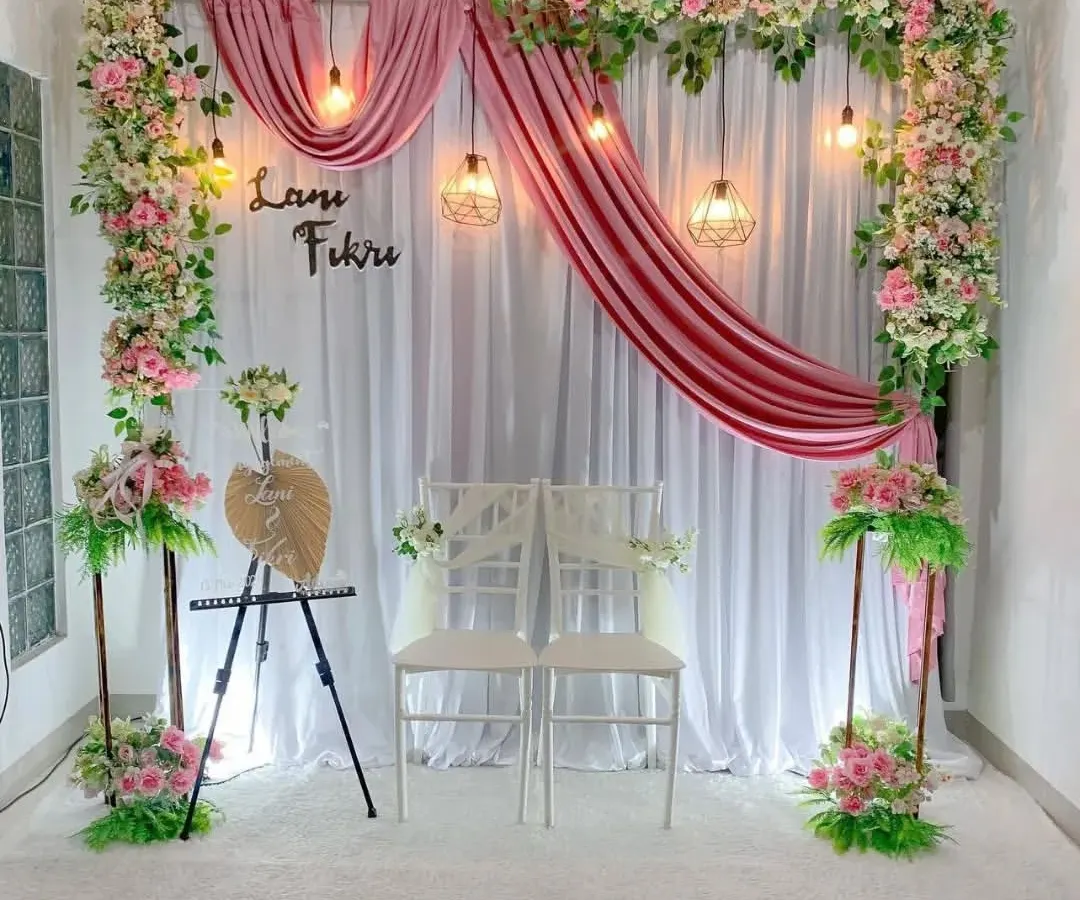 An elegant and romantic engagement stage decoration for an indoor ceremony. This setup includes a white fabric backdrop with gracefully draped pink velvet, framed by lush pink and white floral arrangements and illuminated by warm hanging pendant lights.
