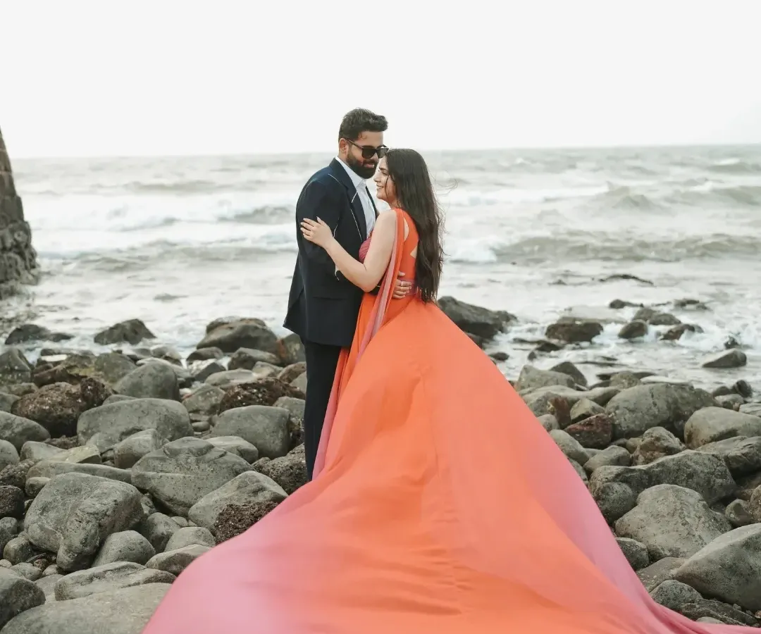 A dramatic shot on a rocky Goa beach, where the bride's vibrant orange gown flows in the wind against the powerful waves, creating a moment of pure cinematic romance.