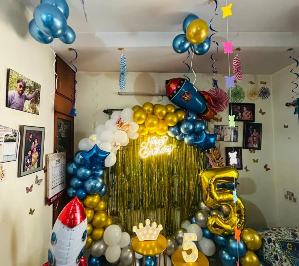 For this 5th birthday, I designed an outer space theme for a celebration at home. The decoration features a large balloon arrangement with rocket ship foil balloons, a gold fringe backdrop, and a rented 'Happy Birthday' neon sign. The large foil number '5' makes the milestone clear.
