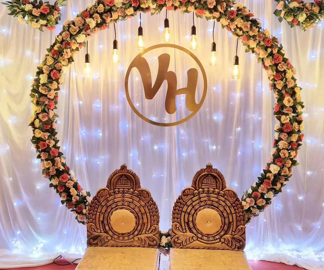 I designed this elegant engagement stage with a large floral ring of artificial roses, warm Edison bulb lighting, and a custom gold monogram for the couple. The traditional chairs complete this beautiful setup for a ring ceremony.