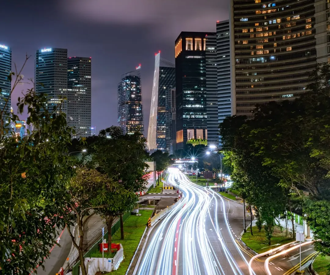 A long-exposure shot of a Singapore highway at night, transforming vehicle headlights and taillights into brilliant white streaks of light against the modern cityscape.