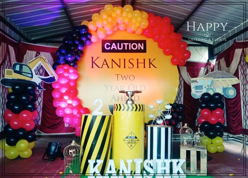 A creative construction theme for a second birthday party. The setup includes a bold "CAUTION" sign on a yellow backdrop, balloon arches in red, yellow, and black, and custom props like themed barrels and vehicle cutouts.
