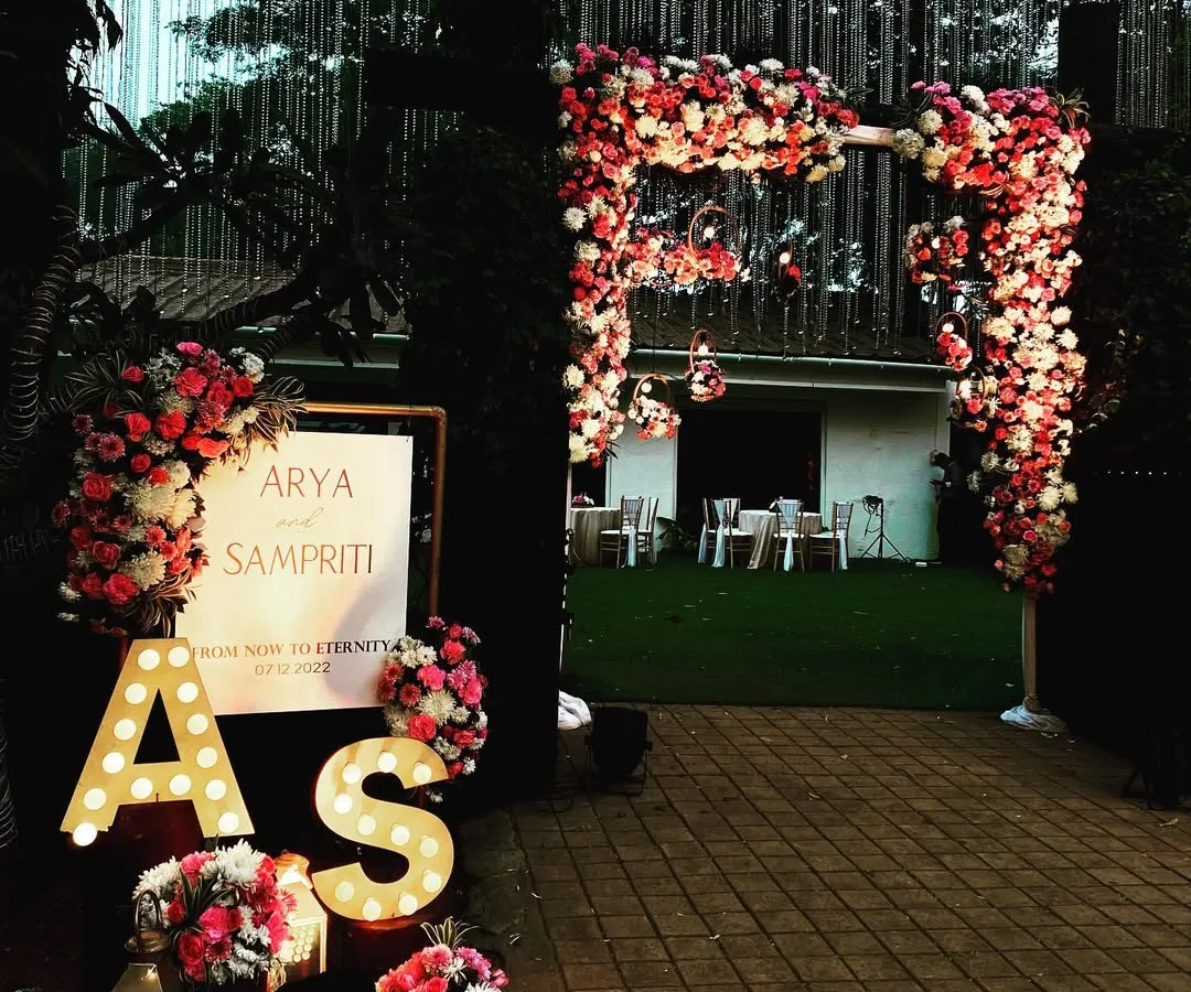 I created this personalized welcome area for an outdoor evening wedding. The entrance features a dense floral arch of pink and white roses, complemented by a custom sign with the couple's initials in marquee lights for a modern and inviting touch.