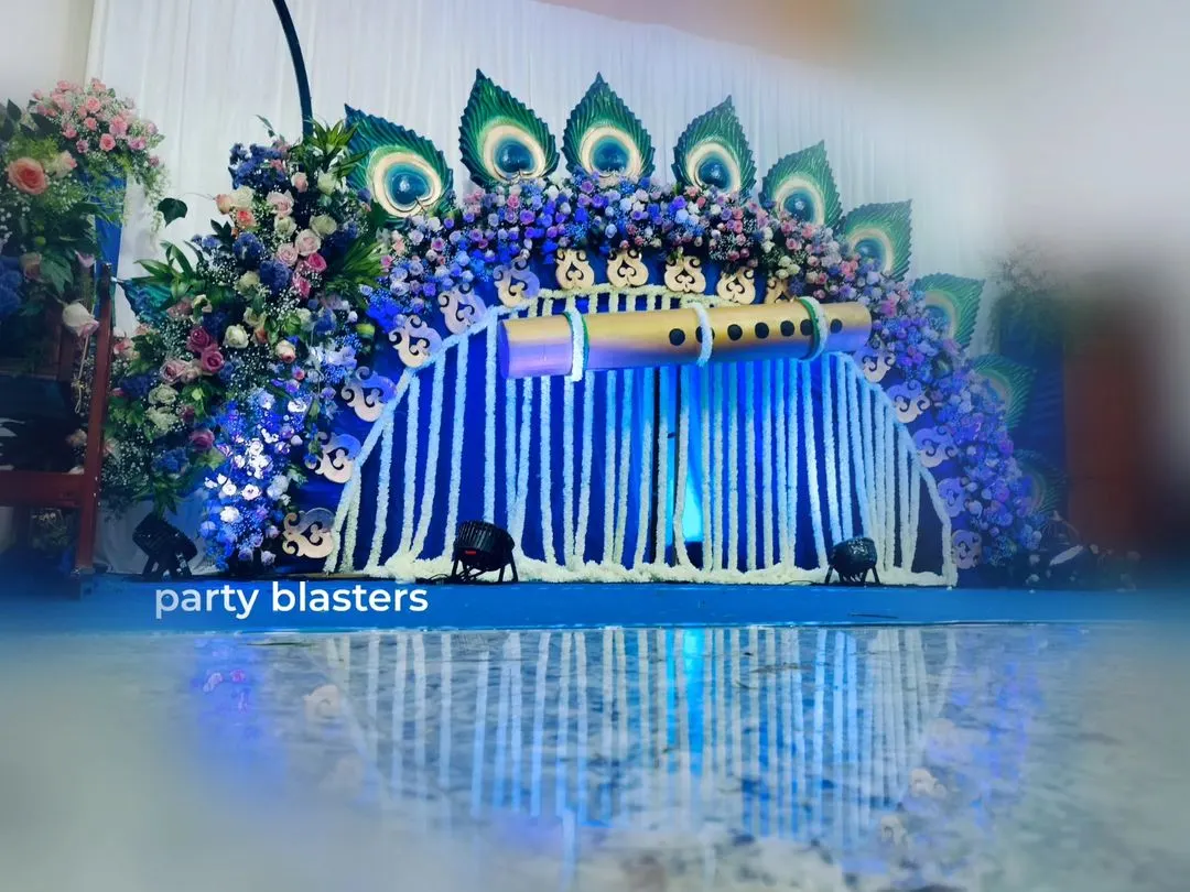 A full view of my popular Krishna-themed naming ceremony decor. The setup features a custom-built backdrop with a large flute, peacock feather details made from natural materials, and a cascade of white tuberose garlands, creating a divine and vibrant setting for the celebration.