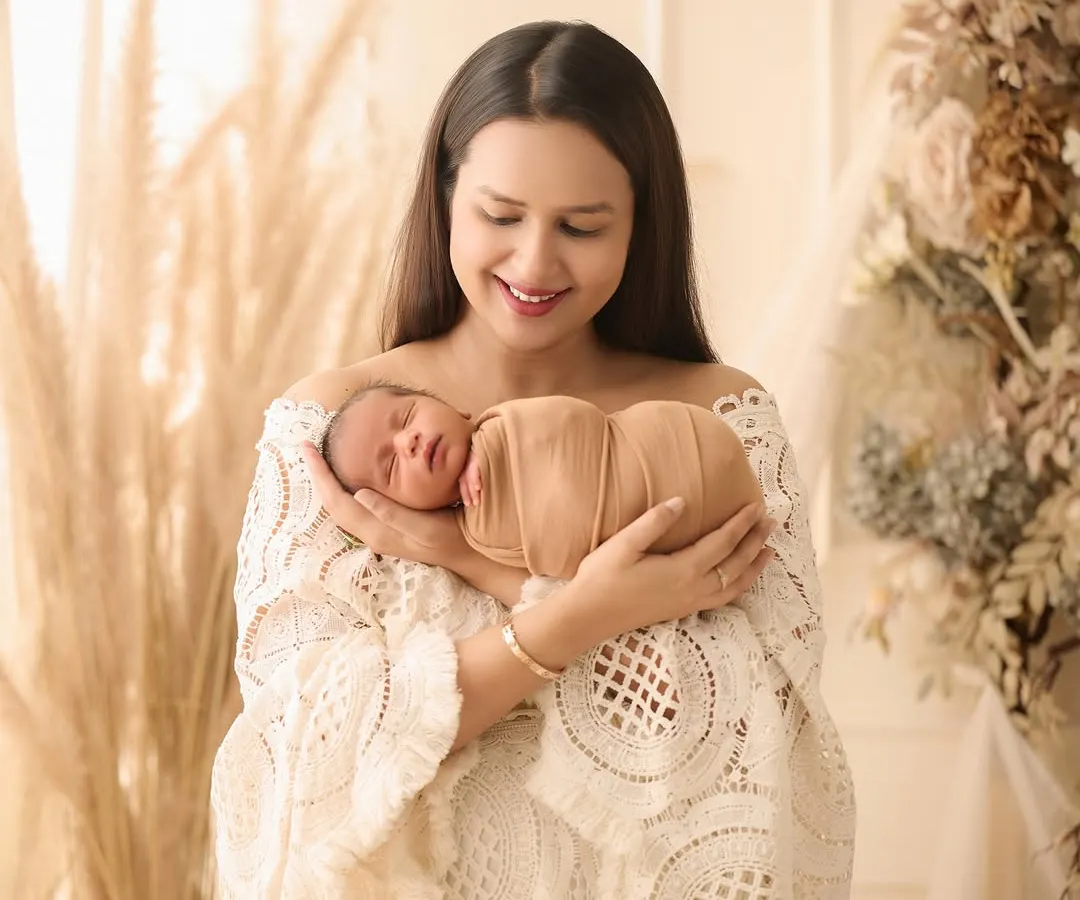 A mother, dressed in a lovely boho-style lace top, lovingly holds her swaddled newborn against a backdrop of pampas grass, capturing a moment of pure tenderness.