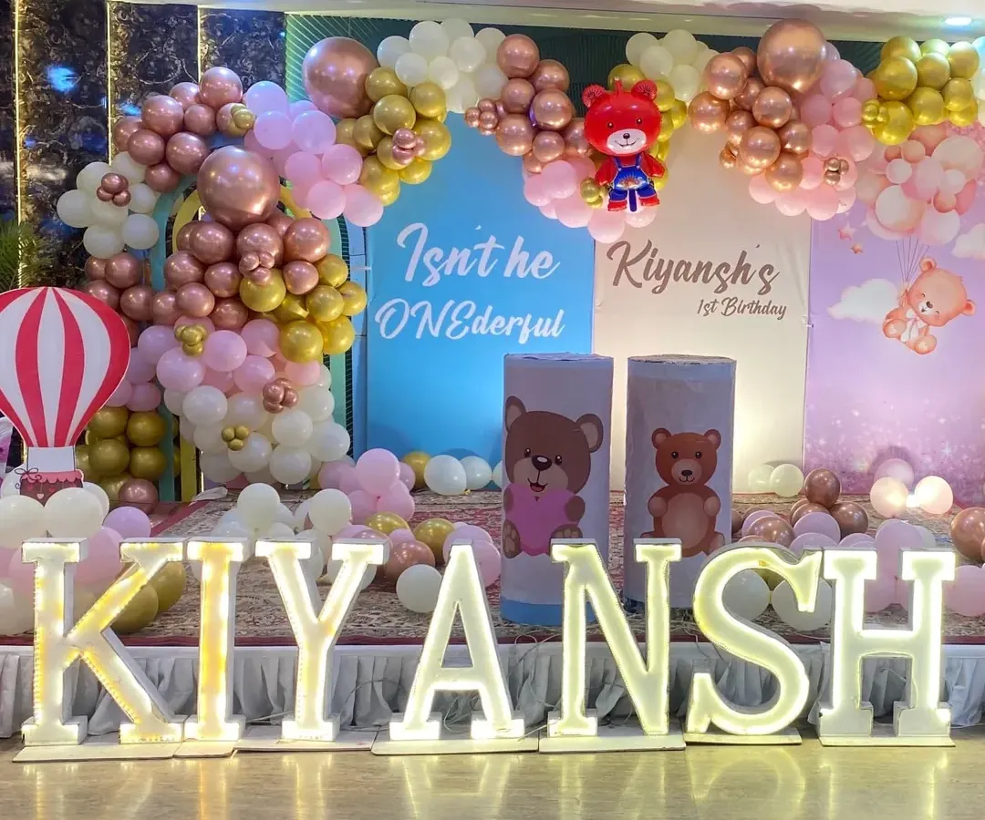 A grand stage setup for a teddy bear themed first birthday party, complete with large marquee letters spelling out the name 'KIYANSH' and multiple themed backdrops.
