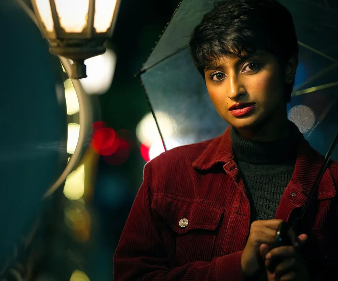 A moody, cinematic portrait of a woman with short hair holding an umbrella under a glowing streetlamp at night. The rich colors and soft focus on the background city lights create a story-driven atmosphere.