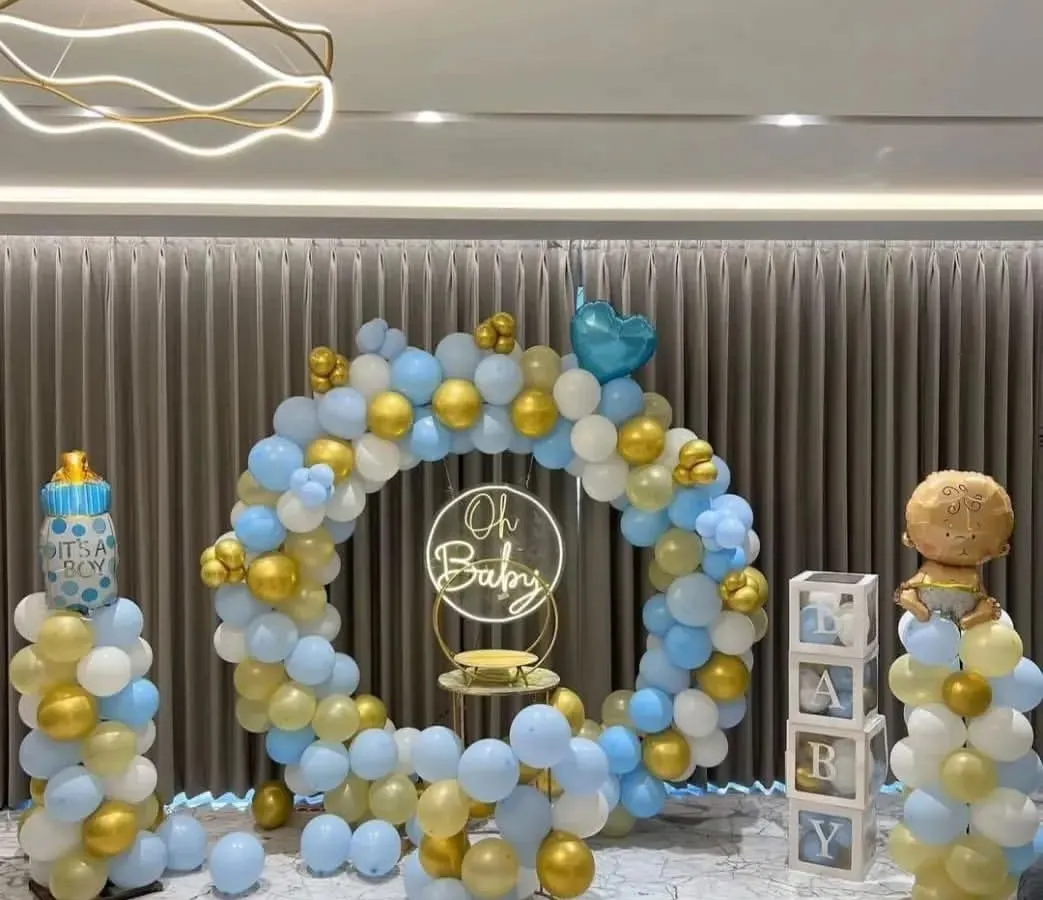 This "Oh Baby" themed baby shower is a classic and elegant choice. The setup features a full balloon ring in a soft blue, white, and gold palette, complemented by a neon sign, "BABY" blocks, and themed balloon pillars.