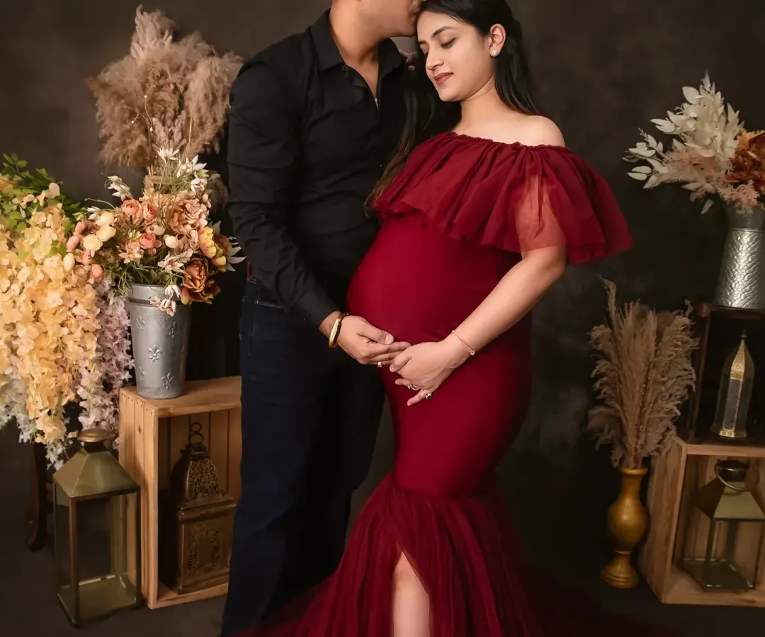 A tender moment between partners. The expecting mother, dressed in a stunning off-the-shoulder red gown, shares a loving glance with her partner, who kisses her forehead in our Gurugram studio.