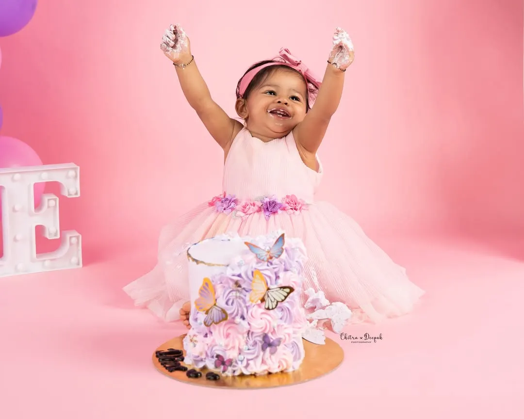 Pure joy and celebration as a little girl, dressed in a lovely pink frock, throws her hands up in delight during her butterfly-themed first birthday cakesmash. These sessions are all about capturing personality and fun.