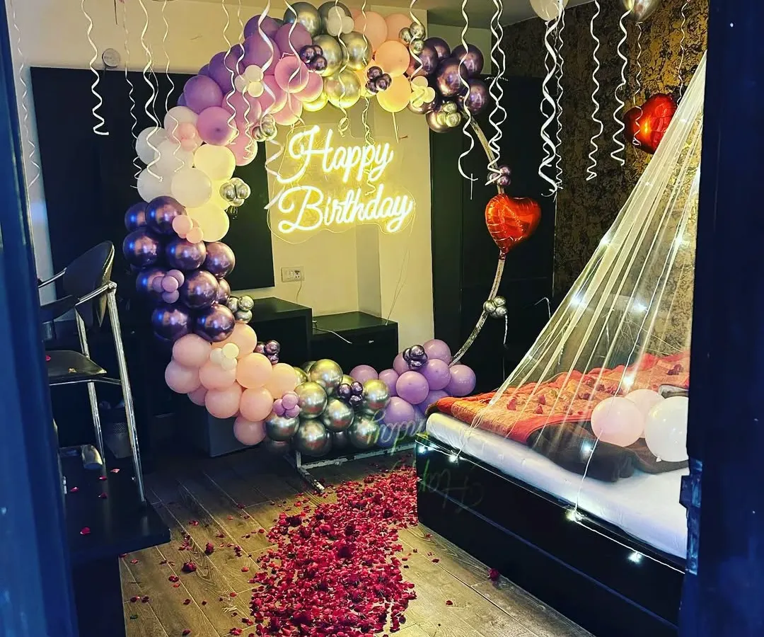 For a truly romantic birthday surprise, I transformed this room with a dreamy setup. It features a beautiful crescent moon balloon arch in shades of purple and pink, a 'Happy Birthday' neon sign, and a delicate canopy over the bed with fairy lights. The floor covered in rose petals completes the magical look.