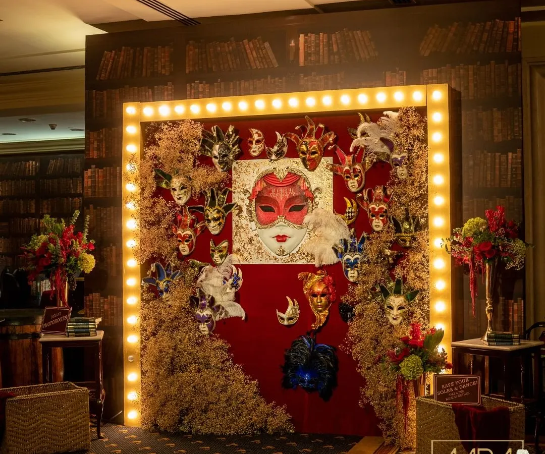 This grand entrance was designed to transport guests to a hidden world of mystery and glamour. The speakeasy masquerade theme came to life with a dramatic photo wall, combining vintage marquee lights with a rich texture of masks and dried foliage against a library-themed backdrop.