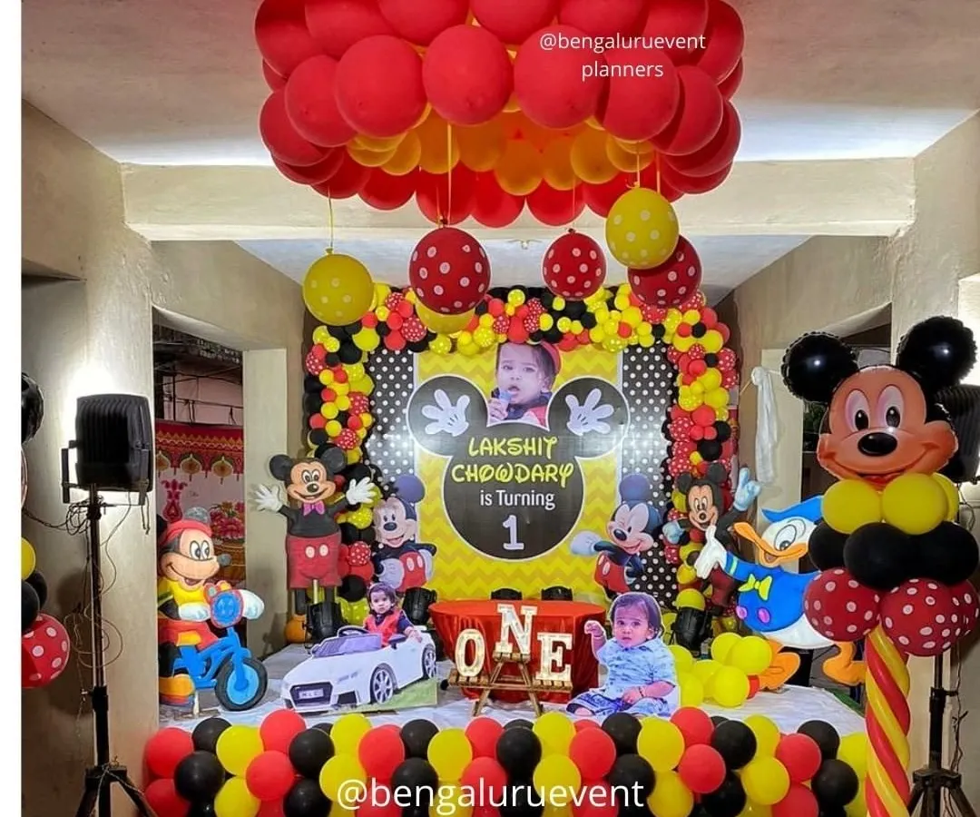 This is a complete Mickey Mouse themed first birthday party I created. The setup includes a personalized backdrop with the child's photo, multiple character cutouts, and coordinated red, yellow, and black balloon decorations for a fun and immersive experience.