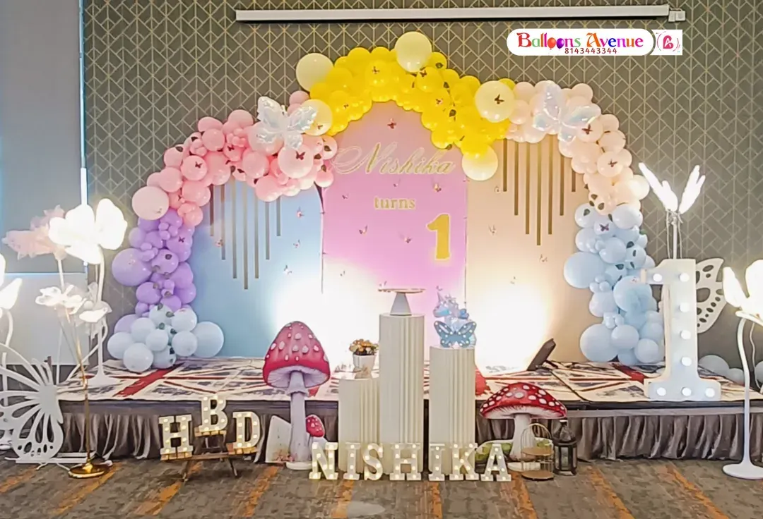We love creating magical first birthday parties like this butterfly garden theme. The setup includes a full stage with a pastel rainbow balloon arch, custom name backdrop, illuminated number one, and whimsical props like giant mushrooms and flower lights.