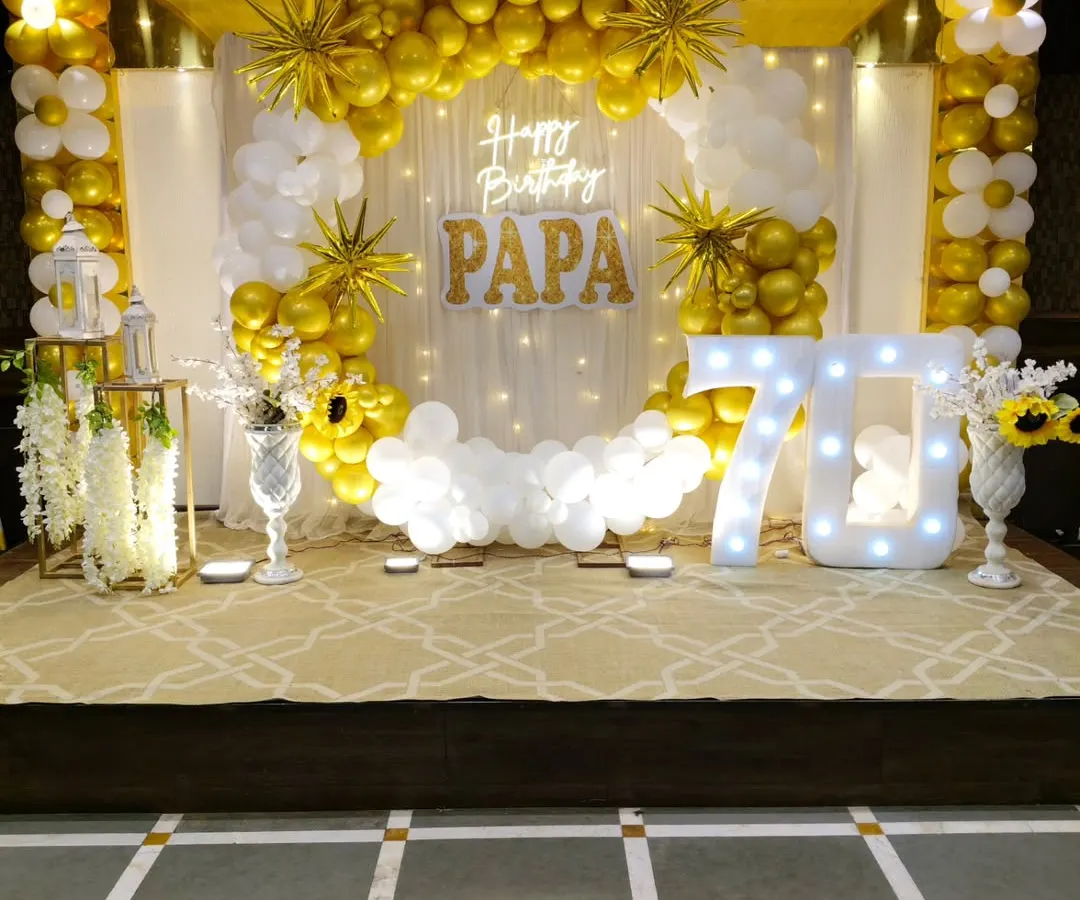 We create elegant setups for milestone celebrations, like this 70th birthday party. The decor features a classic gold and white balloon arch, a sheer curtain backdrop with fairy lights, a custom neon sign, and a large "70" marquee number, creating a sophisticated and celebratory atmosphere.