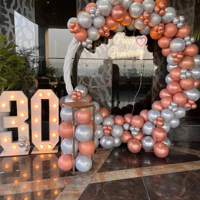 Celebrating a 30th anniversary with a stunning balloon arch against a city skyline. This setup includes a large circular arch in rose gold and silver chrome balloons, a custom 'Happy Anniversary' neon sign, and large light-up marquee numbers for the milestone year.