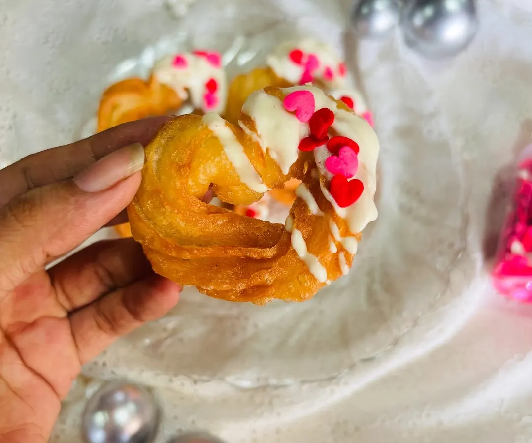 Here is a close-up of my signature heart-shaped churro. You can see the crispy, golden texture of the churro, generously drizzled with creamy white chocolate and topped with red and pink heart-shaped sprinkles for a festive touch.