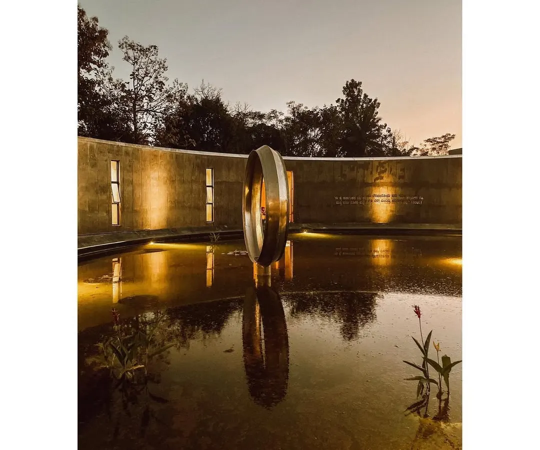 As dusk settles, the Dr. Vishnuvardhan Memorial Complex transforms. The central water body reflects the warm illumination on the concrete walls and the metallic ring sculpture, creating a serene atmosphere for evening contemplation. This is a space designed to honor memory through tranquility.