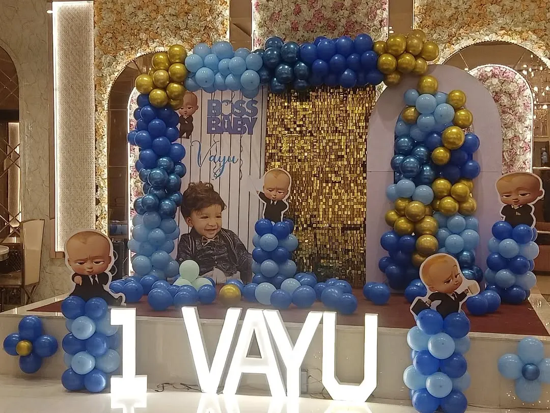 The Boss Baby theme is a huge hit for first birthdays. Here is a complete stage setup for Vayu's party in Noida, featuring a custom backdrop with his photo, character cutouts, and a blue and gold balloon arch, all tied together with large "1 VAYU" marquee letters.