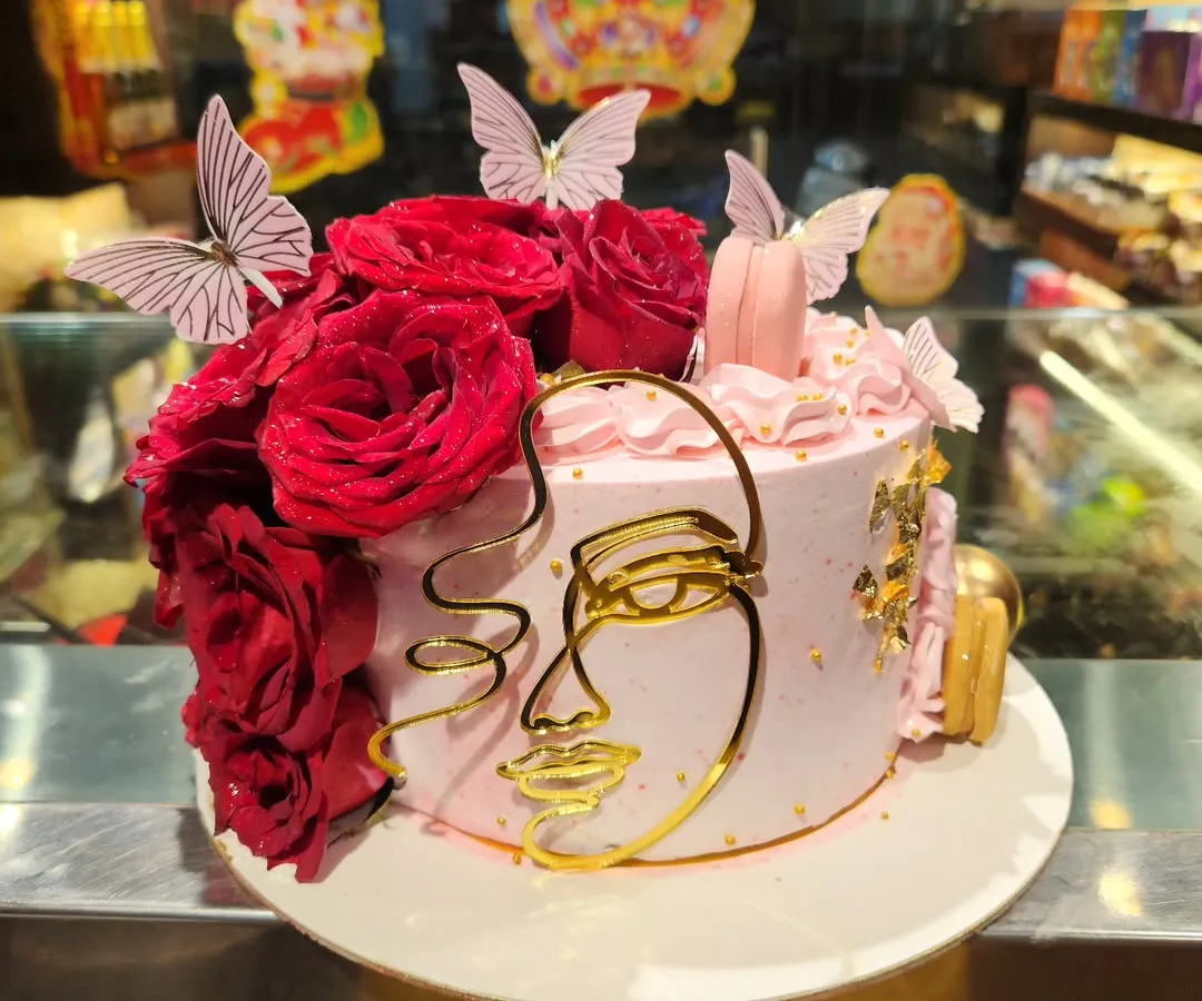 An artistic and modern cake design for a sophisticated celebration. This piece features a delicate pink buttercream finish, a cascade of fresh red roses, edible butterflies, and a striking gold abstract face line art topper, perfect for an elegant birthday or event.