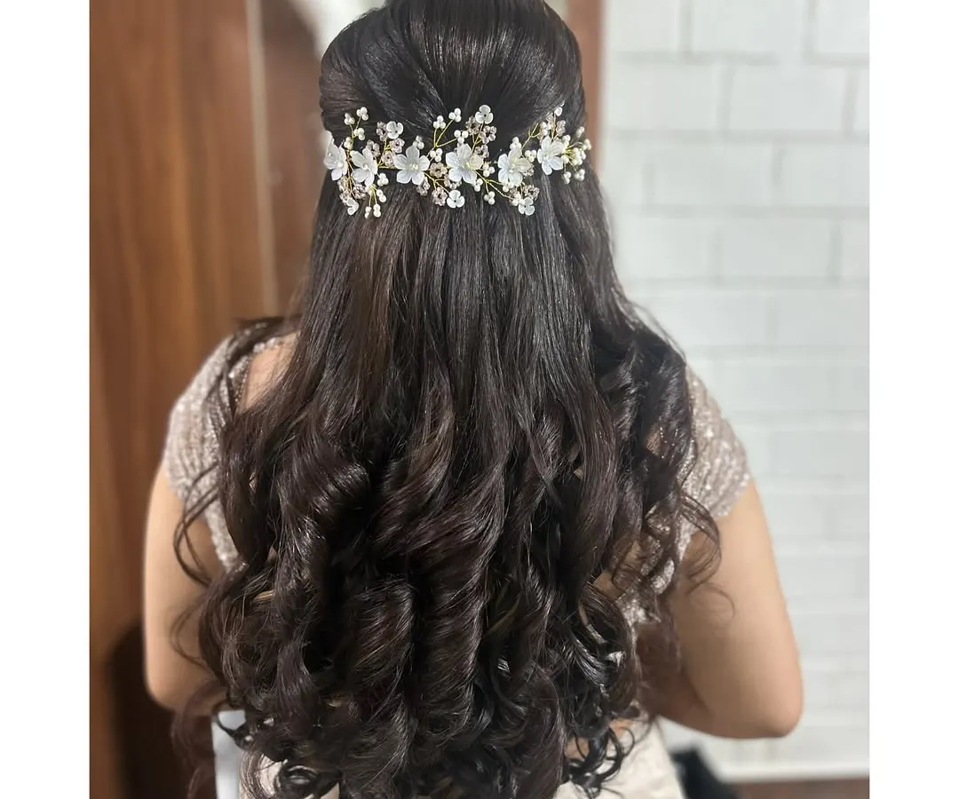 A perfect hairstyle for a reception or engagement. This is a classic half-up style with voluminous, bouncy curls and a delicate floral hair accessory that adds a touch of modern elegance.