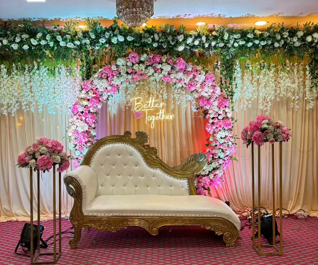 For an elegant engagement, anniversary, or pre-wedding party, we design stunning stage setups. This one features a beautiful floral ring with a 'Better Together' neon sign, complemented by a classic chaise lounge and matching flower stands, creating a sophisticated focal point in a banquet hall.