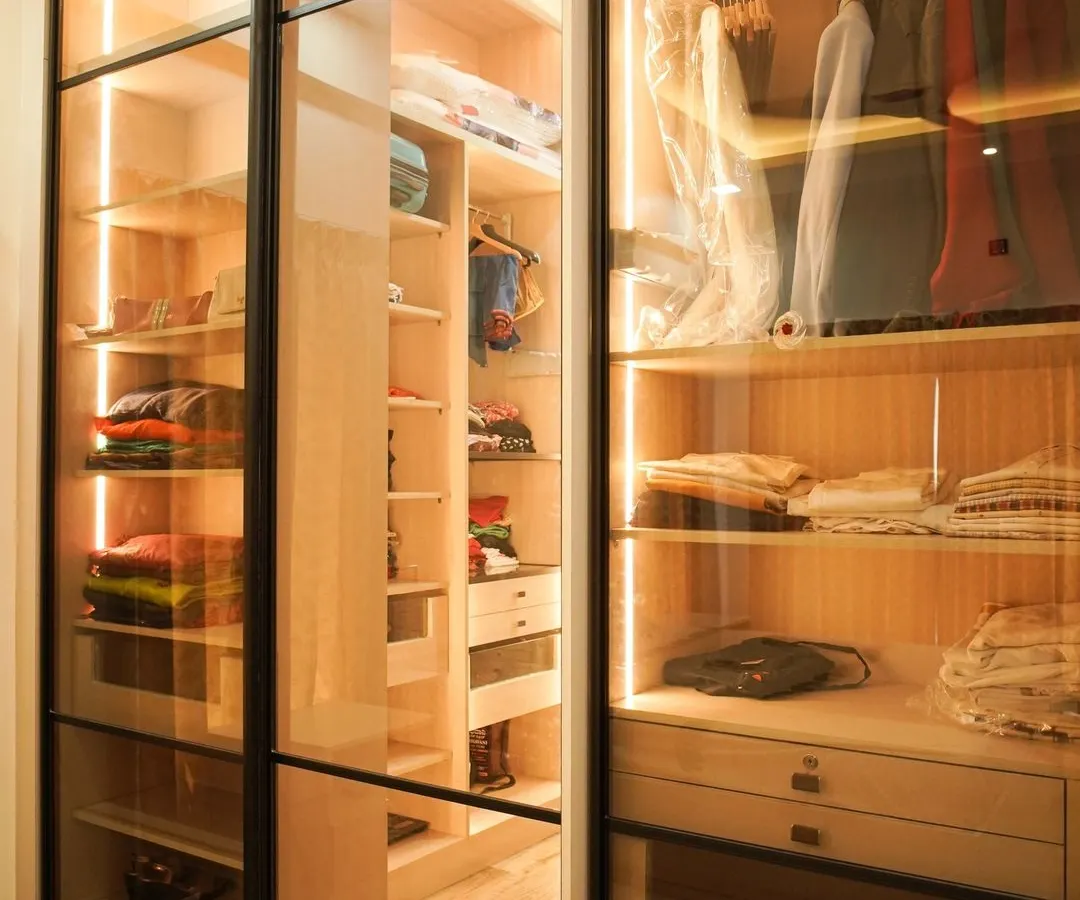 A glimpse into a custom walk-in closet, where luxury meets organization. This design features transparent glass doors, integrated LED lighting, and ample shelving to display and store your collection, turning your wardrobe into a personal boutique.