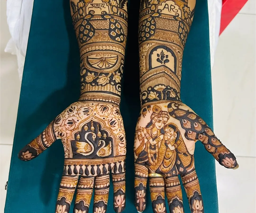 A full bridal mehendi set that showcases a mix of styles. One hand features a beautiful Radha-Krishna figure, while the other has elegant swan motifs and the couple's initials. The arms are decorated with traditional architectural and floral patterns.