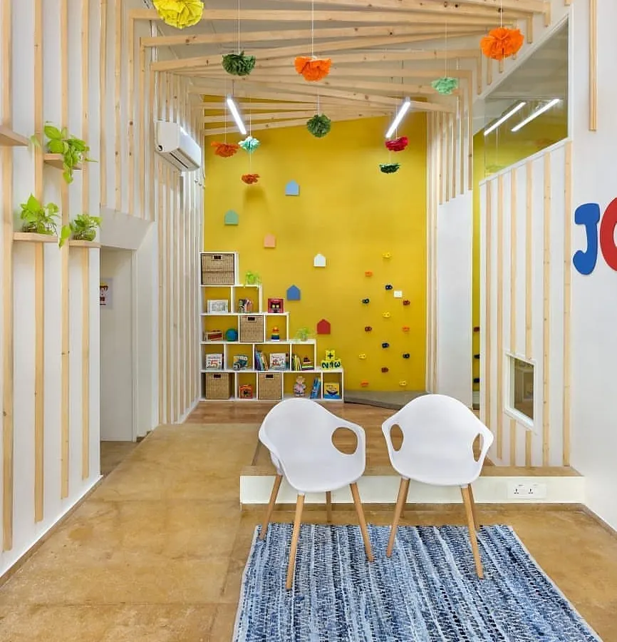 The multipurpose entrance lobby of JOY Preschool, which serves as both a reception area and a space for reading sessions. The design features natural wood slats, custom child-safe furniture, and a small climbing wall against a vibrant yellow backdrop to create an engaging and playful atmosphere.